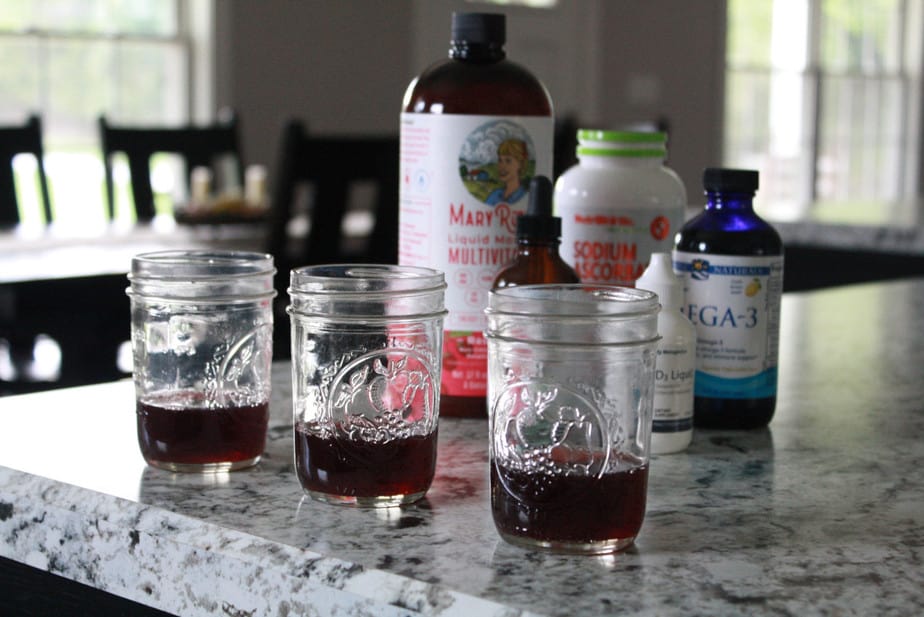 3 cups lined up on kitchen counter with vitamins and grape juice poured in them with the vitamins in the background.