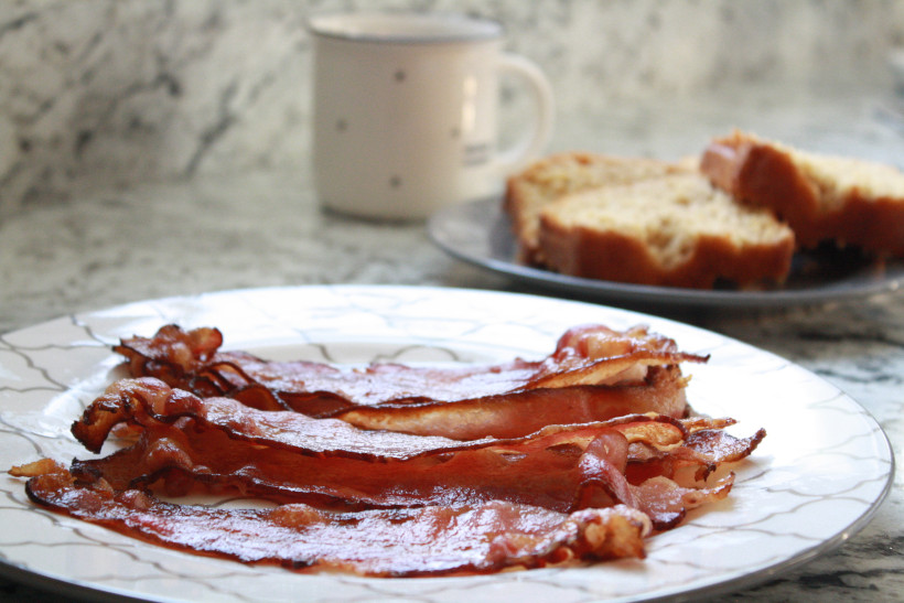 Plate of bacon on kitchen counter with bread and mug in background.