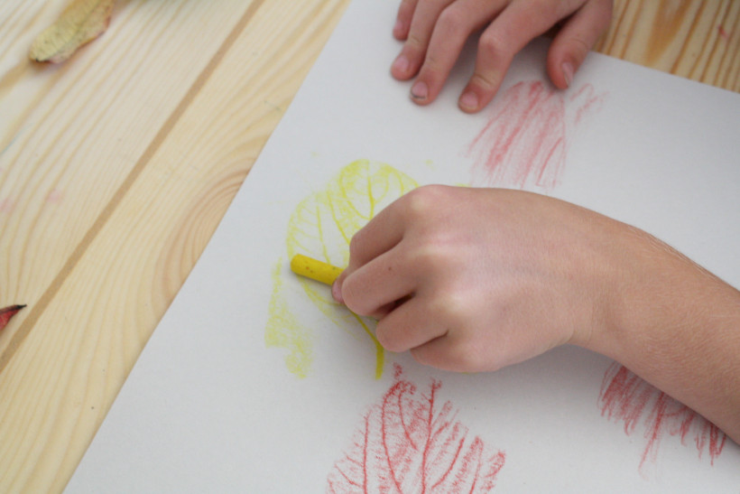 Child using yellow crayon at desk to make a leaf rubbing on white paper.