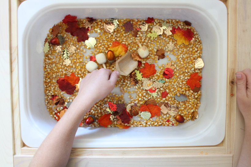 Child playing in sensory bin using an ikea sensory table with popcorn kernels, autumn foil confetti, wooden pumpkins, acorns, and leaf confetti.