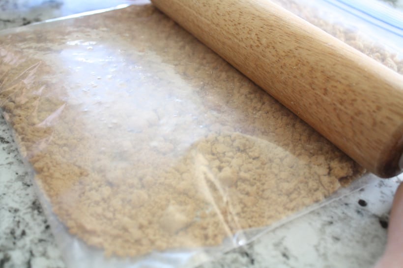 Woman using a rolling pin to crush gingersnap cookies in a plastic bag on kitchen counter.