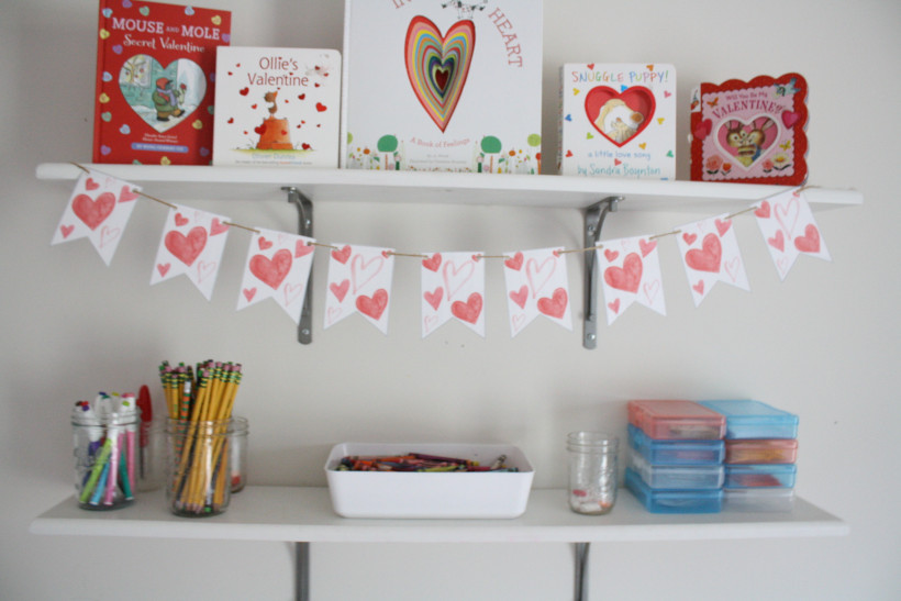 Red and pink free printable Valentine's Day banner displayed on white shelf with Valentine's Day books on above shelf.