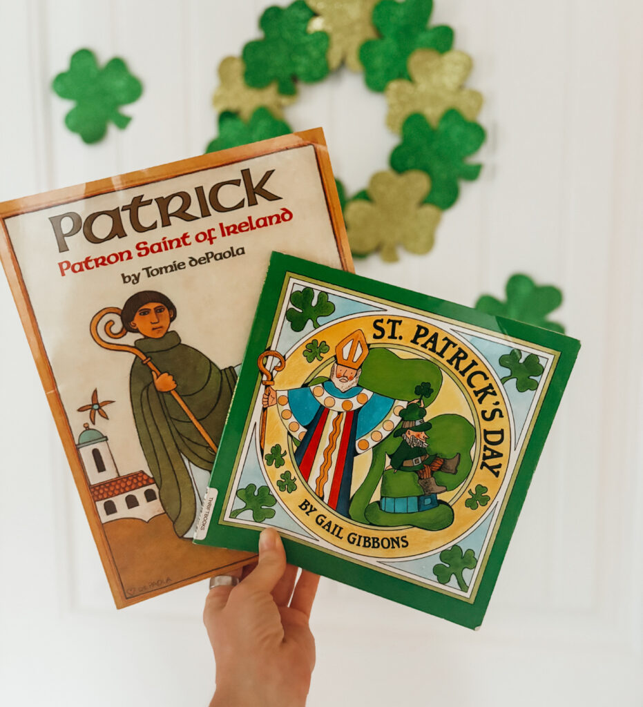 Woman holding 2 of March picture books for kids. One is St. Patrick Patron Saint of Ireland and St. Patrick's Day, in front of white door with green clover wreath.