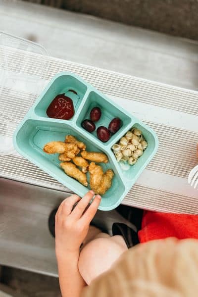Blue bento box with chicken, ketchup, grapes, and mac n cheese at ballpark for summer routines for kids and mom.